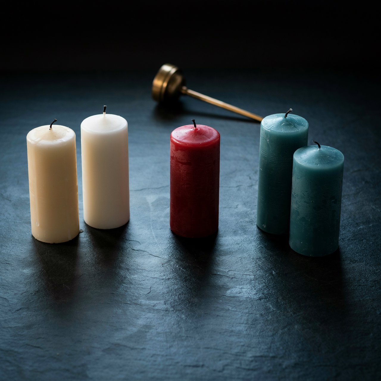 Exactly five candles stand on a dark slate tabletop: two ivory candles on the left, one red candle in the center, and two teal candles on the right. A brass snuffer sits above the red candle, nothing else. — Ideogram v4