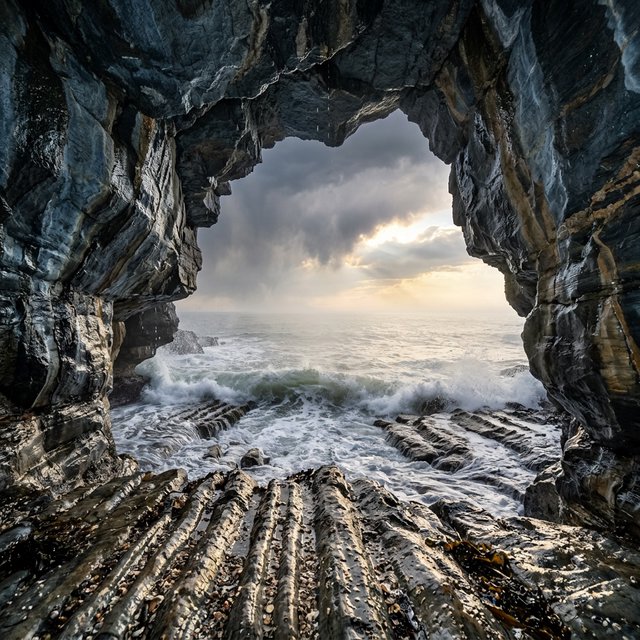 Inside a blue-grey sea cave, incoming swells curl over ribbed limestone beneath a clearing rain squall in late afternoon; wet mineral bands and shell fragments sharpen the entrance ledge, while the cavern mouth frames hazy open water. — Nano Banana 2.1