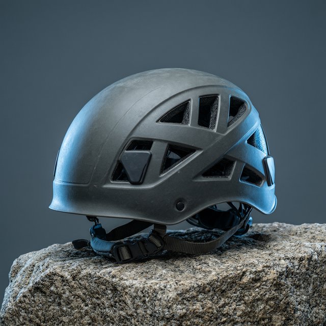 Against a muted slate backdrop, an unbranded climbing helmet with a matte recycled-polymer shell and woven nylon straps rests on rough granite; broad diffused key light and a cool rim light shape this commercial product image, with no lettering. — Qwen Image 3.0 Pro