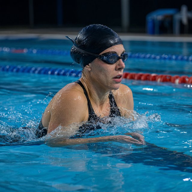 Under the blue-hour floodlights, an adult swimmer drives off the starting block into an outdoor pool. A real-world sports press photo, shot at 200mm with shallow depth of field, captures beads of water suspended around her shoulder. — Qwen Image 3.0 Pro