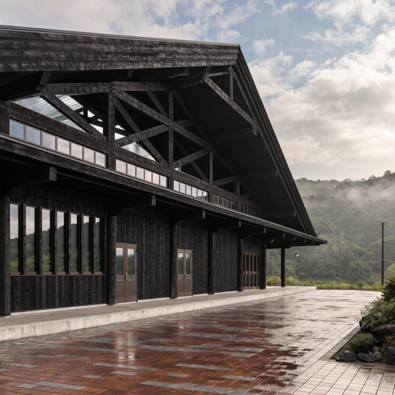 Contemporary Japanese timber train station, charred cedar cladding, pale concrete platform, broad pitched roof with exposed beams and translucent clerestories, cool morning light, viewed obliquely from the quiet approach plaza, precise straight lines and plausible structure. — Recraft v4 Pro