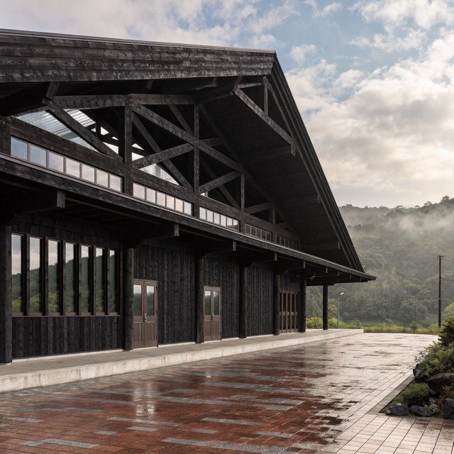 Contemporary Japanese timber train station, charred cedar cladding, pale concrete platform, broad pitched roof with exposed beams and translucent clerestories, cool morning light, viewed obliquely from the quiet approach plaza, precise straight lines and plausible structure. — Recraft v4 Pro