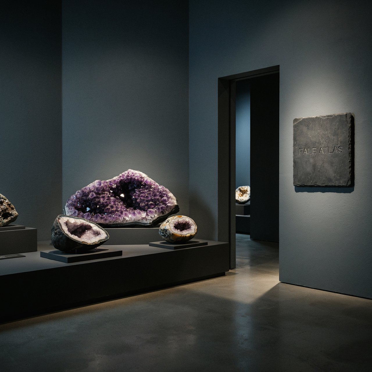Within a quiet mineral gallery, amethyst clusters and polished geodes catch pools of light; "PALE ATLAS" is carved in clean capitals across a slate plaque beside the entrance. — Ideogram v4.5