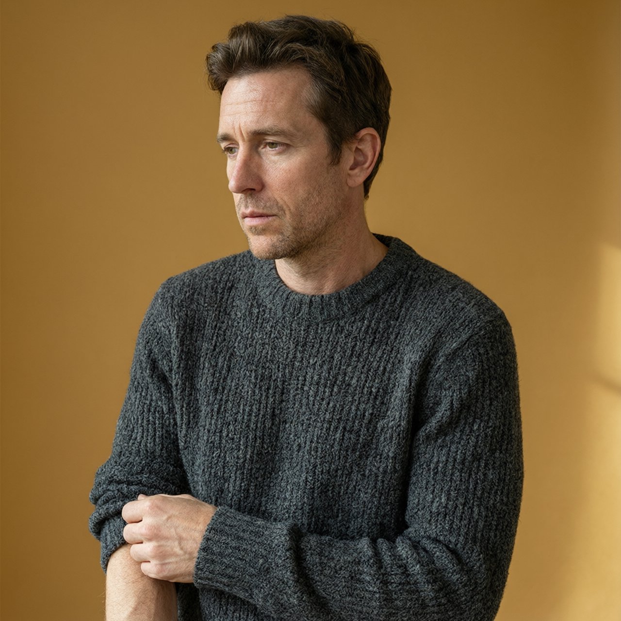 Editorial portrait of an adult man in his early 40s wearing a charcoal wool fisherman’s sweater, one hand loosely holding its ribbed cuff. His thoughtful, slightly tired expression rests against a muted ochre studio backdrop, shaped by gentle directional window light. — Qwen Image 3.0 Pro
