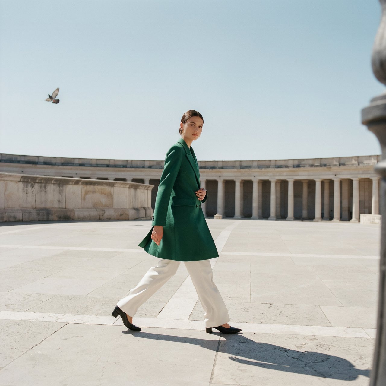 Full-length fashion editorial: model in a structured emerald wool coat and white trousers walking across a wide empty plaza, harsh midday sun, long shadow. — Ideogram v4