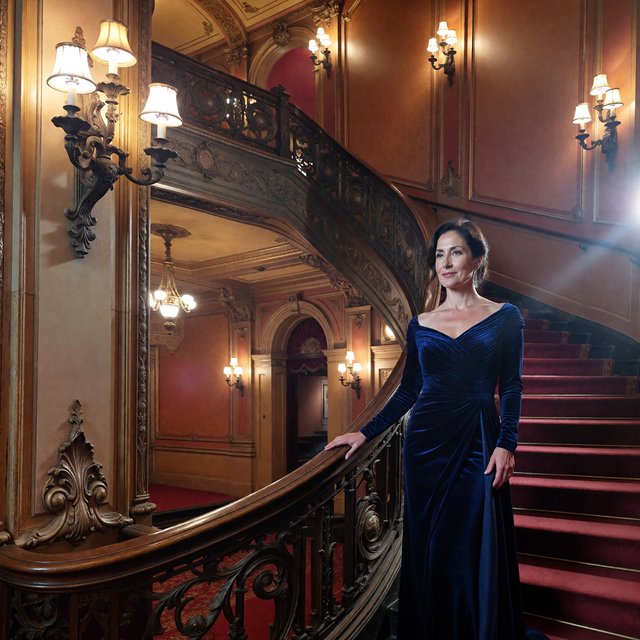Editorial glamour photograph of an adult woman in a midnight-blue velvet evening gown, standing naturally beside a curved staircase in an old theatre, warm flattering sconce light, relaxed shoulders, poised expression, refined cinematic elegance. — ImagineArt 1.5 Pro
