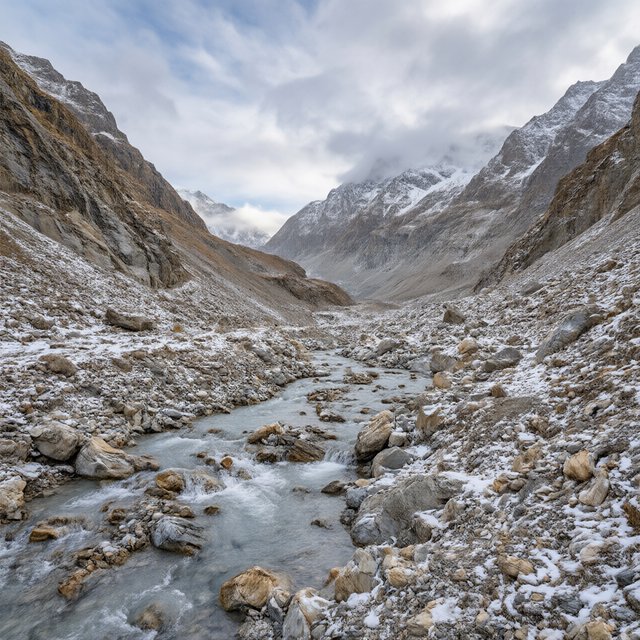 Beyond a braided glacier-fed stream, a steep moraine valley opens beneath clearing dawn snow flurries; gritty ice grains and water-polished stones sharpen the near bank, while layered ridges fade into cloud. — Model 17
