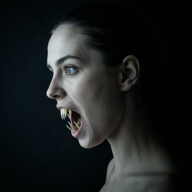 A creepy woman in side profile. Focus on the head with a slightly alien figure. Pale skin. No irises in the eyes. Mouth is open wide with very sharp, long razor teeth. Black background. Chiaroscuro effect. — Ideogram v4.5