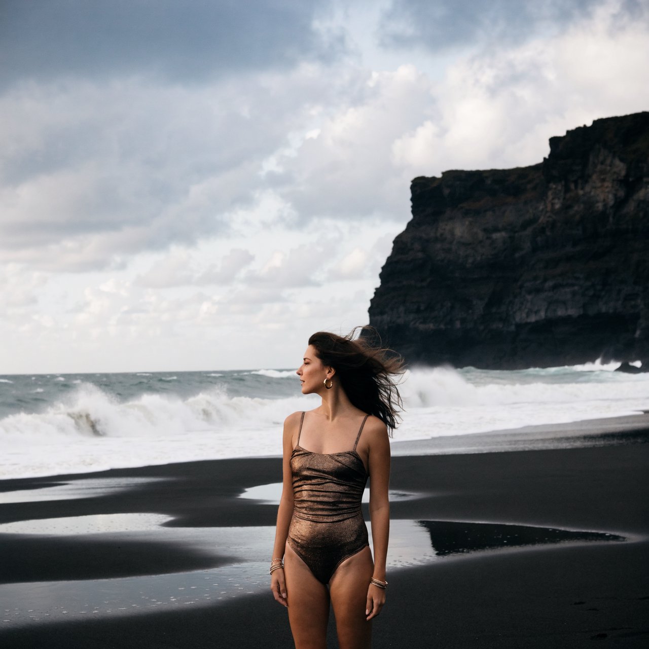 Editorial glamour photograph of an adult woman in a bronze one-piece swimsuit, standing at ease on a quiet black-sand beach beside tide pools, flattering overcast light softly shaping the scene, wind-lifted hair, natural gaze toward distant surf. — Recraft v4 Pro
