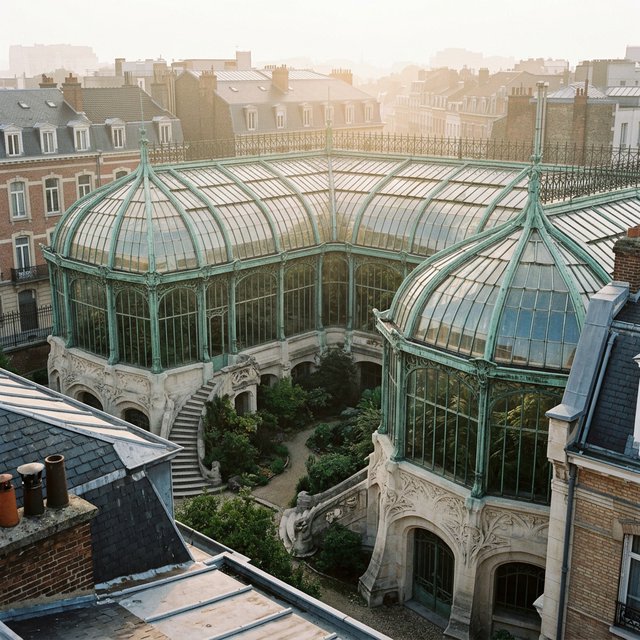 From a nearby rooftop, an Art Nouveau conservatory curves around a planted courtyard, its green copper ribs, clear glass, and pale stone base catching hazy morning light; orderly rooflines and believable supports show from above. — Nano Banana 2.1