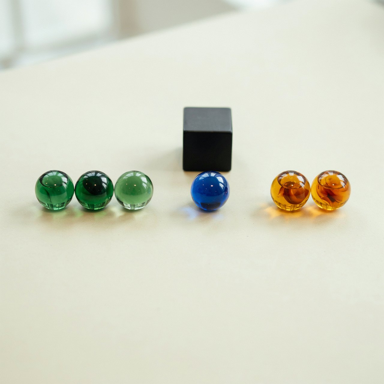 Exactly six glass marbles sit on a cream tabletop: three green marbles in a row on the left, two amber marbles in a row on the right, and one blue marble centered between the rows. A black cube sits above the blue marble, nothing else. — Ideogram v4