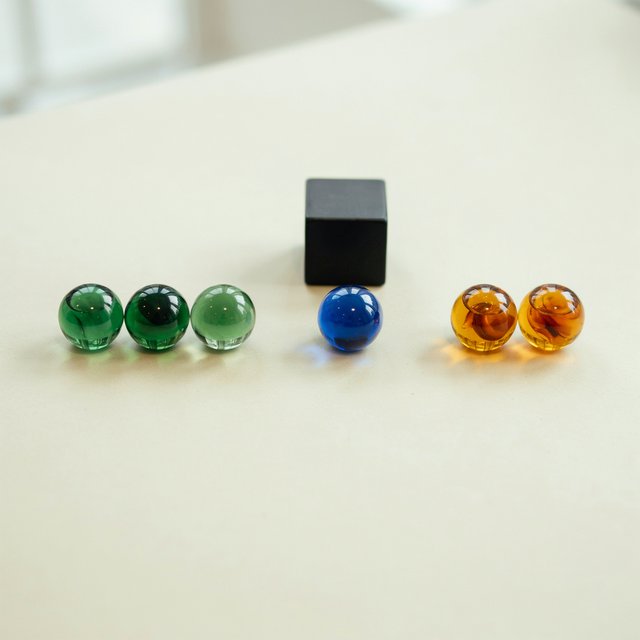 Exactly six glass marbles sit on a cream tabletop: three green marbles in a row on the left, two amber marbles in a row on the right, and one blue marble centered between the rows. A black cube sits above the blue marble, nothing else. — Ideogram v4