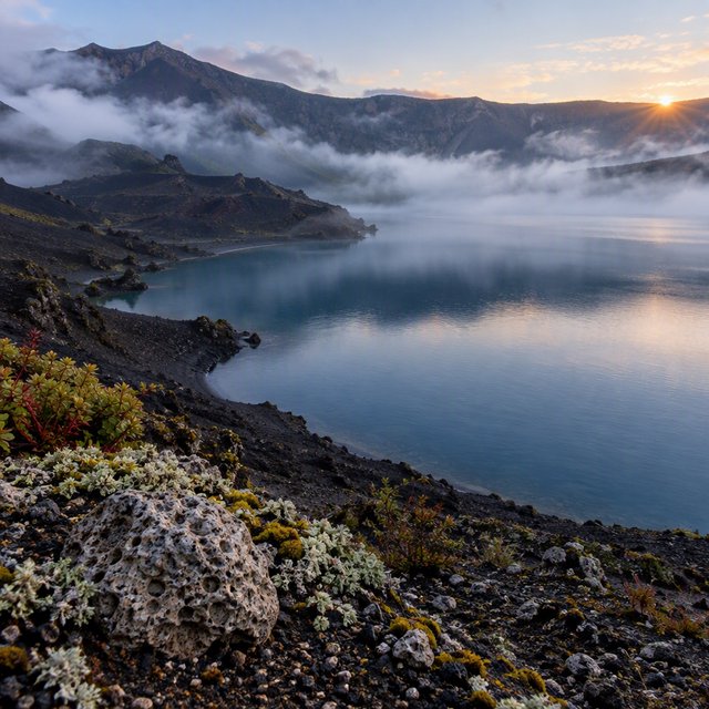 At first light, a rain-cleared volcanic caldera lake lies beneath thinning fog; pumice grains and damp lichen sharpen the near shore, while dark cinder slopes step back toward a distant rim. — GPT Image 2.5 Flare