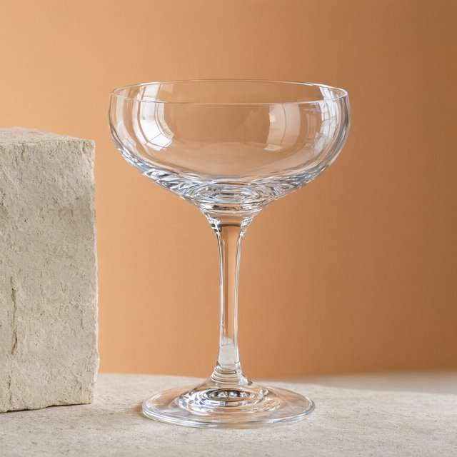 Beside a pale limestone block, an unbranded handblown cocktail coupe in clear soda-lime glass catches crisp window-shaped highlights; its curved bowl bends a muted apricot backdrop under broad diffused key light and narrow cool rim lighting, commercial product image, no lettering. — Qwen Image 3.0 Pro
