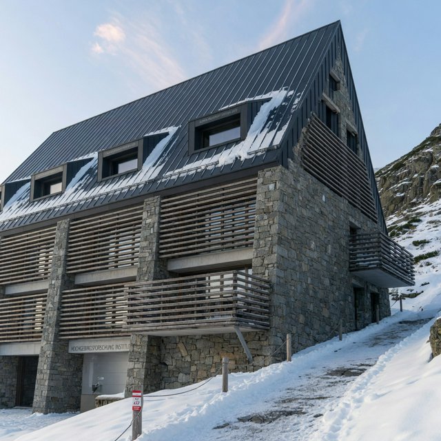 Exterior of a contemporary alpine research lodge in restrained regional modernism, rough granite walls, larch screens, steep standing-seam roof, crisp snow-bright morning light, viewed from below across a mountain footpath, sound structural geometry. — Grok Imagine Image Quality