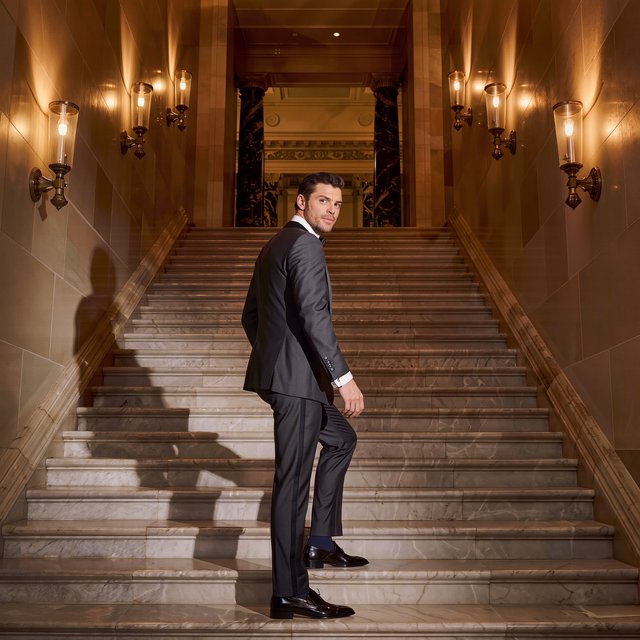 A tall adult man in a charcoal evening suit pauses halfway down a sweeping hotel staircase, caught glancing toward the ballroom as amber sconces cast sculpted light across stone; crisp tailoring and composed fashion-magazine polish. — Model 9