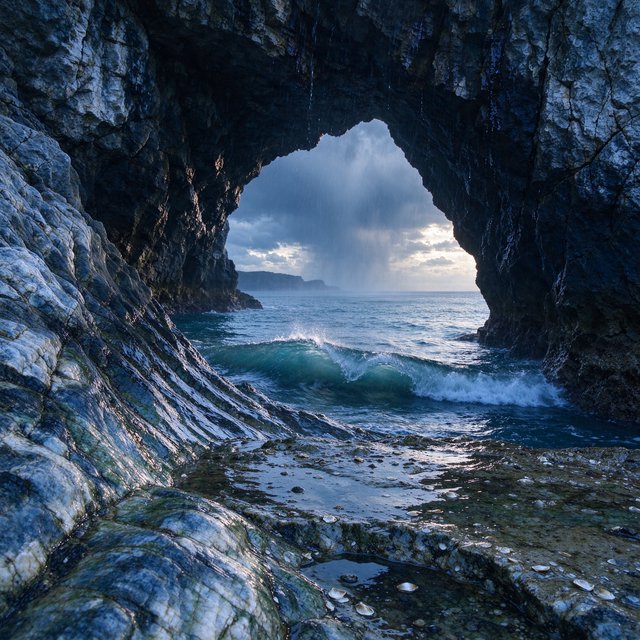 Inside a blue-grey sea cave, incoming swells curl over ribbed limestone beneath a clearing rain squall in late afternoon; wet mineral bands and shell fragments sharpen the entrance ledge, while the cavern mouth frames hazy open water. — Seedream 5.0 Flash