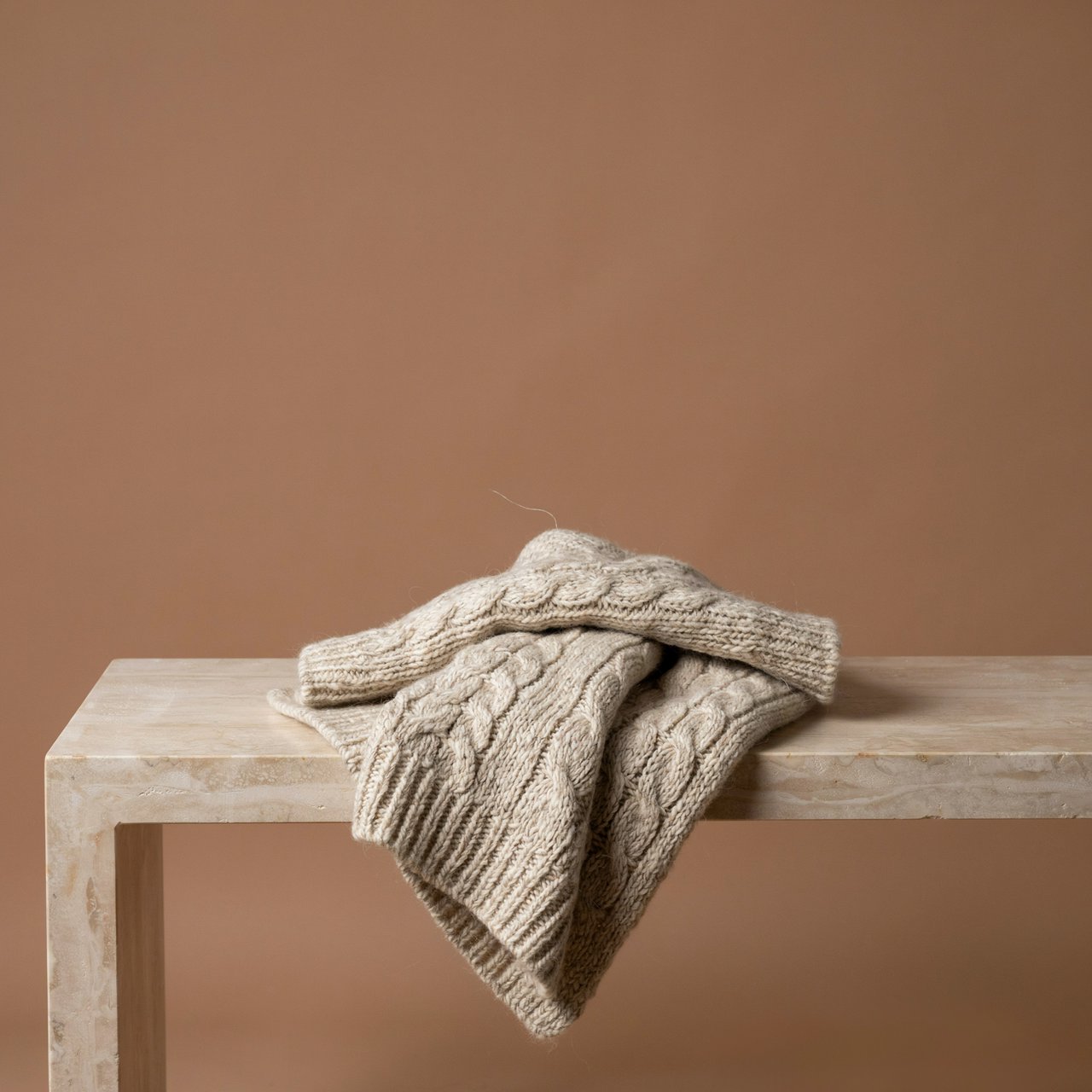 Soft folds of an unbranded chunky-knit wool throw rest on a pale travertine bench; its looped surface catches broad diffused key light and gentle side fill against a muted terracotta studio background, forming a clean commercial product image with no lettering. — Ideogram v4