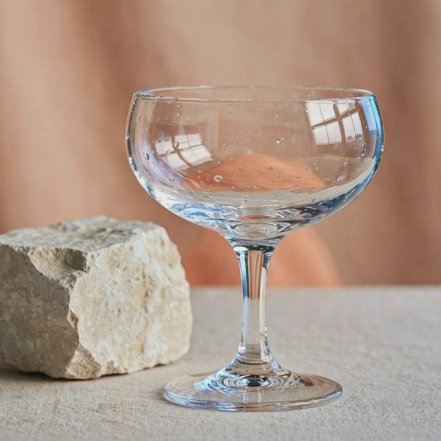 Beside a pale limestone block, an unbranded handblown cocktail coupe in clear soda-lime glass catches crisp window-shaped highlights; its curved bowl bends a muted apricot backdrop under broad diffused key light and narrow cool rim lighting, commercial product image, no lettering. — Muse Image 1.0
