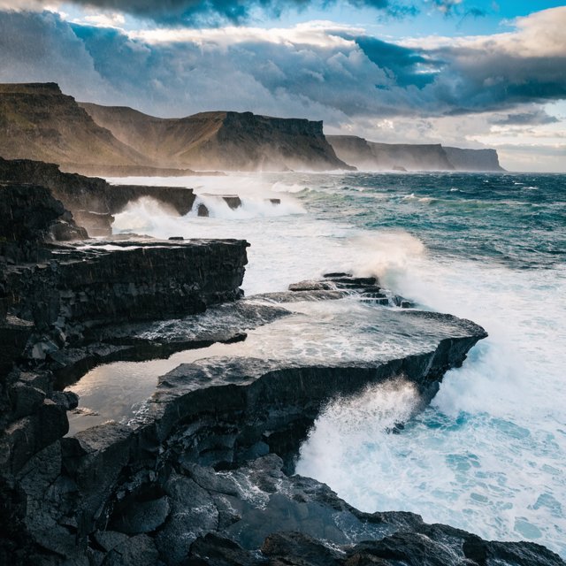 A rugged volcanic coastline under clearing storm clouds at late afternoon, black basalt shelves descending into turquoise surf, sea spray clinging to jagged rocks, tide pools reflecting distant cliffs and layered mountains fading into rain haze. — Recraft v4