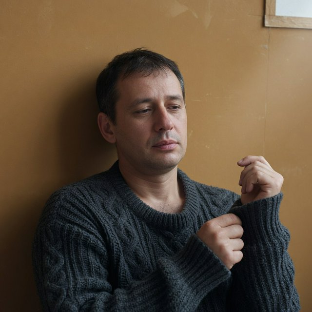 Editorial portrait of an adult man in his early 40s wearing a charcoal wool fisherman’s sweater, one hand loosely holding its ribbed cuff. His thoughtful, slightly tired expression rests against a muted ochre studio backdrop, shaped by gentle directional window light. — Grok Imagine Image Quality