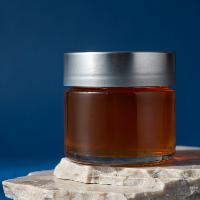 A squat unbranded honey jar with clear glass walls and a plain brushed-aluminum lid rests on honed pale limestone; broad diffused key light and warm rim light shape the commercial product image against a seamless cobalt-blue background, with no lettering. — Model 8