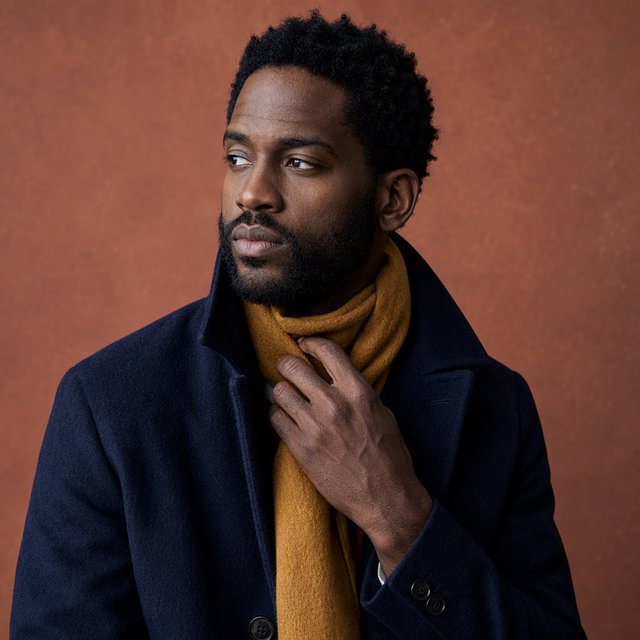 Editorial portrait of an adult Black man in his 30s wearing a deep navy wool overcoat and mustard scarf, fingers adjusting the scarf, thoughtful expression, muted terracotta studio backdrop, crisp cool window light. — GPT Image 2
