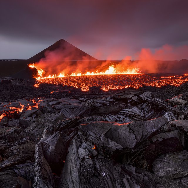 Fresh lava threads a black cinder plain on a volcanic island at dusk beneath ash-laden wind; blistered crust and brittle pumice in the foreground give way to glowing fissures fading into smoke. — Kling v3