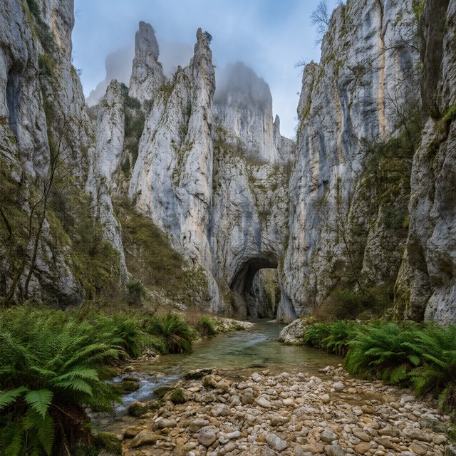 A limestone karst valley at blue hour after a light shower, towering pale cliffs rising above a clear river, low clouds snagging on pinnacles, slick ferns and rounded pebbles leading toward a distant stone arch. — Qwen Image 3.0