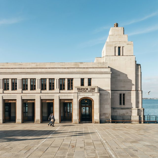 Exterior of a coastal Art Deco ferry terminal, cream limestone, dark bronze window frames, stepped geometric tower and sheltered arcade, late-morning sunlight casting sharp shadows, viewed from across a windswept stone forecourt, realistic architectural photography with precise lines and plausible structure. — Ideogram v4