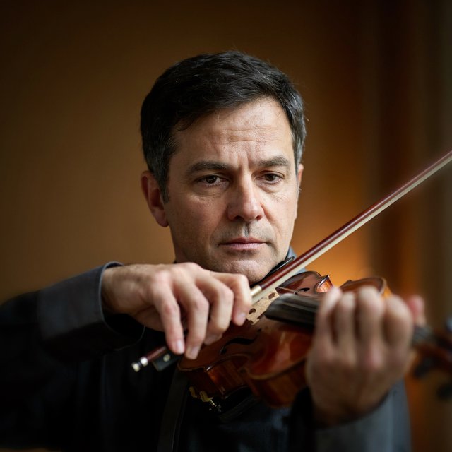 With a violin tucked close, an adult man in his early 50s draws a bow, his hands prominent, wearing a charcoal silk shirt; his intent expression is framed against a warm umber backdrop in soft amber window light, in a close portrait photograph. — Ideogram v4.5
