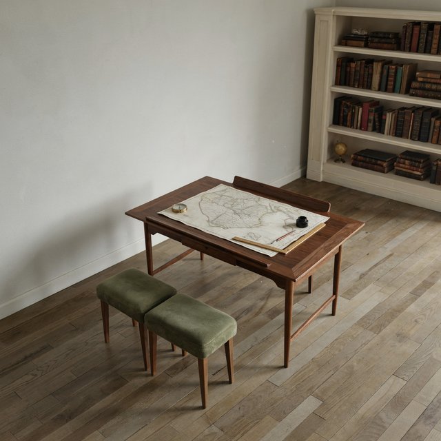 A quiet mapmaking room contains exactly one walnut drafting desk at the center, two moss-green stools side by side along its west edge, and one cream bookcase against the north wall; pale morning light, nothing else. — Ideogram v4
