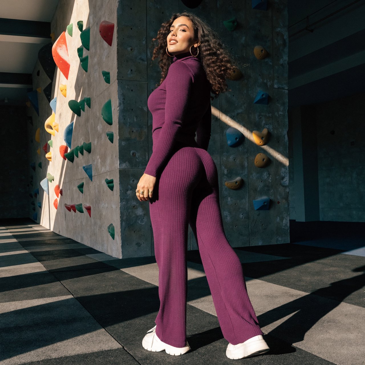 Editorial glamour photograph of an adult woman in a plum fitness set, pausing naturally beside a sunlit indoor climbing wall, flattering side light tracing colorful holds, loose curls and an easy, confident smile. — Recraft v4 Pro
