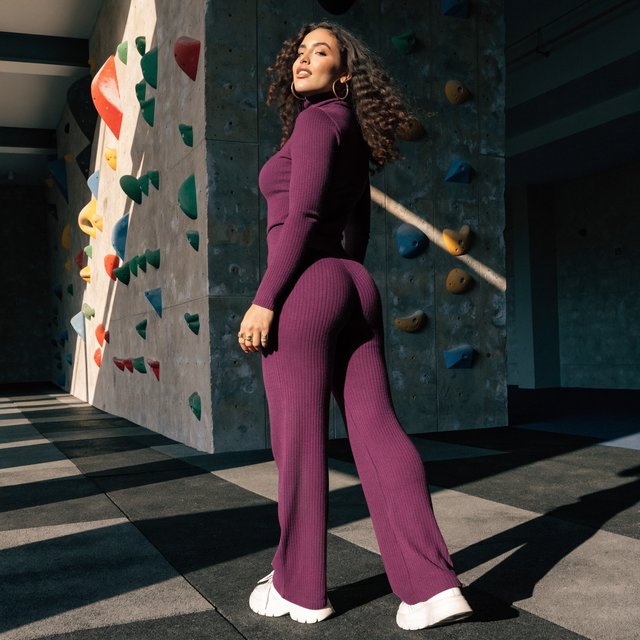 Editorial glamour photograph of an adult woman in a plum fitness set, pausing naturally beside a sunlit indoor climbing wall, flattering side light tracing colorful holds, loose curls and an easy, confident smile. — Recraft v4 Pro