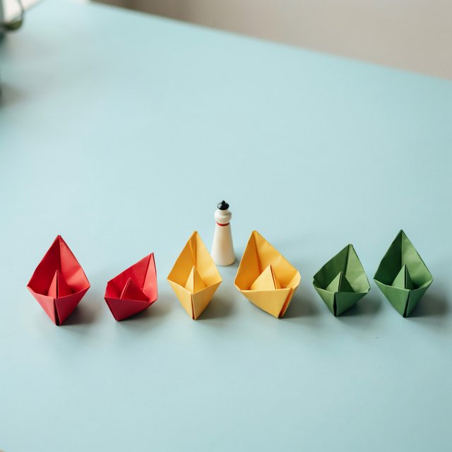 Exactly six folded paper boats sit on a pale blue tabletop: two red boats on the left, two yellow boats in the center, and two green boats on the right. A small white lighthouse figurine stands behind the center pair, nothing else. — Ideogram v4