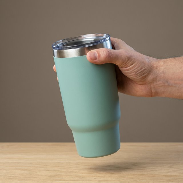 An adult hand lifts an unbranded insulated travel mug, its matte sea-glass polymer body and brushed-steel rim catching softbox highlights above a pale oak counter; warm gray studio backdrop, broad diffused key and cool edge light, no face or lettering. — Qwen Image 3.0 Pro