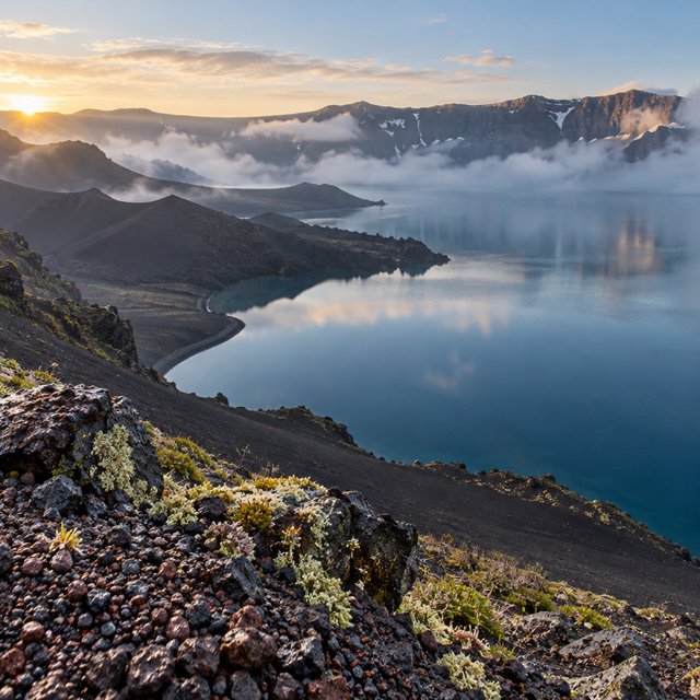 At first light, a rain-cleared volcanic caldera lake lies beneath thinning fog; pumice grains and damp lichen sharpen the near shore, while dark cinder slopes step back toward a distant rim. — GPT Image 2.5 Sunburst
