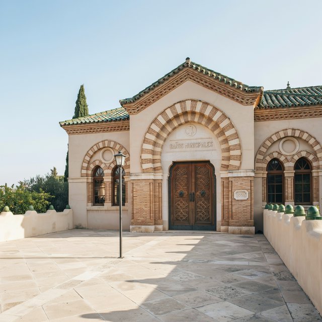 Exterior of a Moorish Revival municipal bathhouse, pale buff stucco, striped brick arches, green glazed tile roof and carved cedar doors; clear morning light, viewed across a paved courtyard, with coherent structure and crisp lines. — Ideogram v4