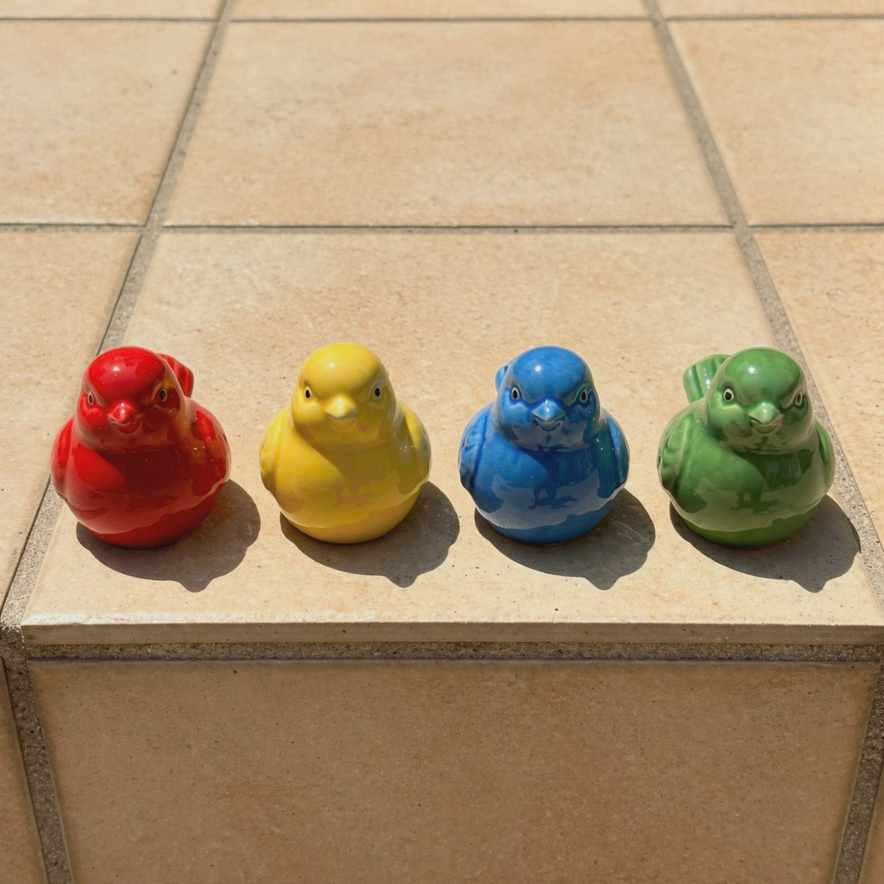 On a sunlit tiled ledge, exactly four ceramic bird figurines form a row: red at far left, yellow at left-center, blue at right-center, and green at far right; all face forward, nothing else. — Muse Image 1.0