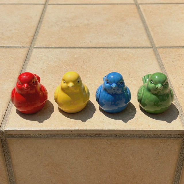 On a sunlit tiled ledge, exactly four ceramic bird figurines form a row: red at far left, yellow at left-center, blue at right-center, and green at far right; all face forward, nothing else. — Muse Image 1.0