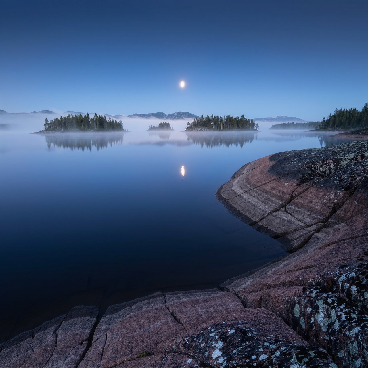 An expansive boreal lake beneath a clear, cold midnight sky, pale moonlight catching thin mist above dark water; spruce-covered islands recede toward distant ridges, while lichen and cracked granite sharpen the near shoreline. — Kling v3 Omni