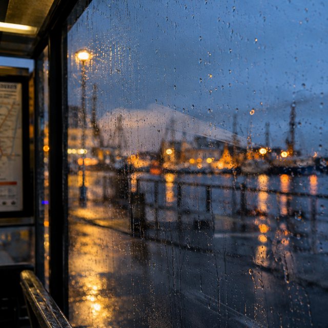 Raindrops crawl down a bus shelter pane, warping blurred harbor lights beyond. A handheld evening street snapshot in cool sodium glow, shot at 50mm with shallow depth of field, catches tiny water trails and a faint wipe mark. — GPT Image 2.5 Flare