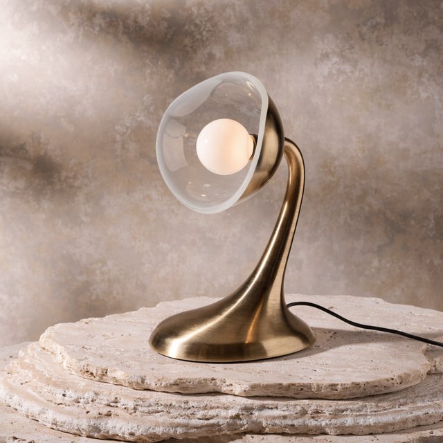 Commercial product photograph of an unbranded sculptural desk lamp made from brushed brass and opal glass, resting on pale travertine, against a warm gray background, with soft diffused light from upper left and gentle grounding shadow. — Recraft v4