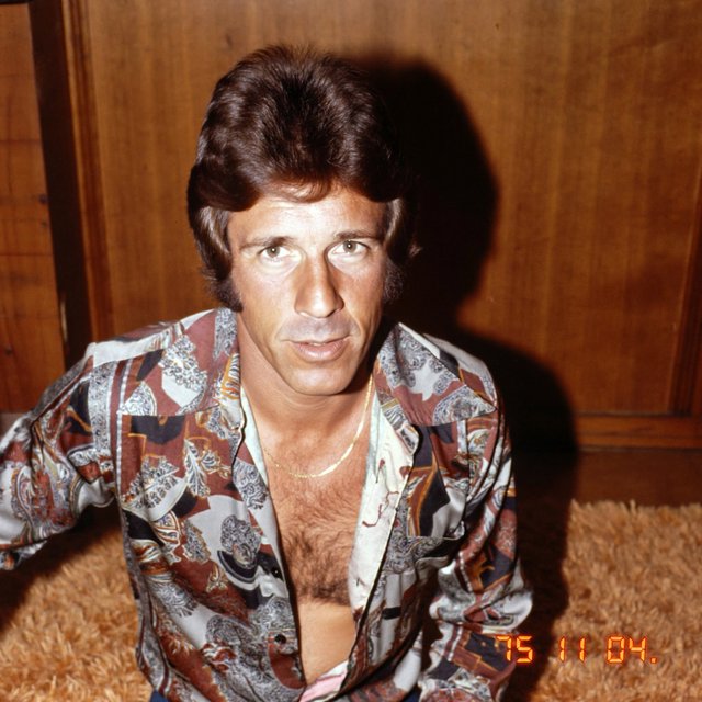 Create an image of a man in the quintessential style of 1975. Style his hair, clothing, accessories, and surroundings to reflect the distinctive look of the mid-1970s—featuring bold silhouettes, eye-catching accessories, layered details, and characteristic colors and textures. Give the image the feel of an authentic 1975 photograph, complete with analog film grain, faded colors, direct flash lighting, and a slight soft-focus effect. Add a period-accurate, red-orange date stamp in a 1970s style in one of the bottom corners. No recognizable posters. No modern objects or text. — ImagineArt 2