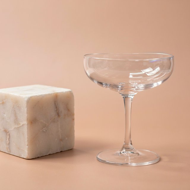 Beside a pale limestone block, an unbranded handblown cocktail coupe in clear soda-lime glass catches crisp window-shaped highlights; its curved bowl bends a muted apricot backdrop under broad diffused key light and narrow cool rim lighting, commercial product image, no lettering. — Ideogram v4.5