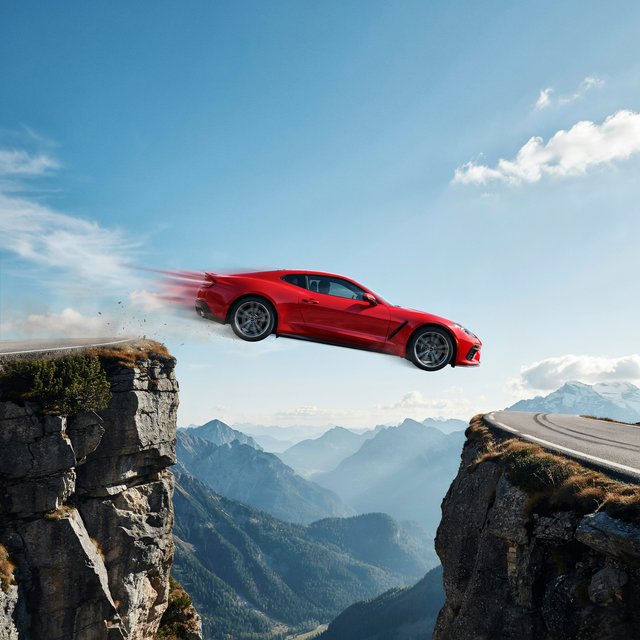 A sports car in a daredevil stunt has driven off a road from a high cliff, attempting to fly and land on the road on the next cliff. Beautiful mountainous background, sunny blue sky. Show motion, speed, and action. — Ideogram v4.5