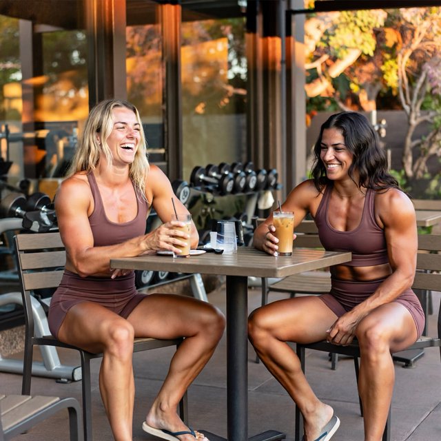 Two muscular adult women in their twenties, one Caucasian blonde and one Latina, sit at an outdoor café table beside a gym after their workout. They wear tight workout shorts, sports bras, and flip-flops. — ImagineArt 2