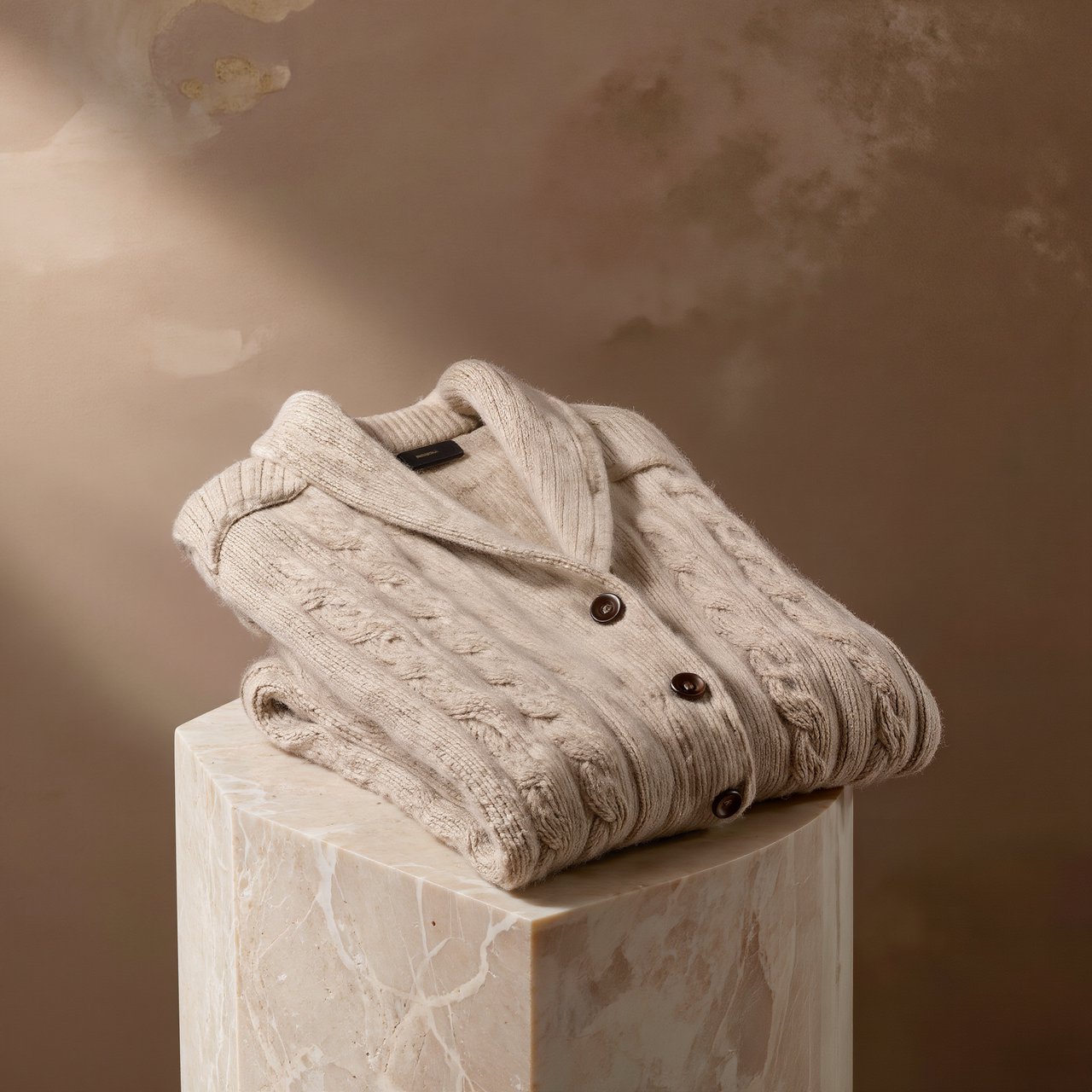 A folded unbranded merino cardigan rests on a honed limestone plinth, its chunky ribbed wool surface softly catching broad overhead diffusion and warm side bounce against a muted clay backdrop, with no tags, lettering, or graphics. — Kling v3