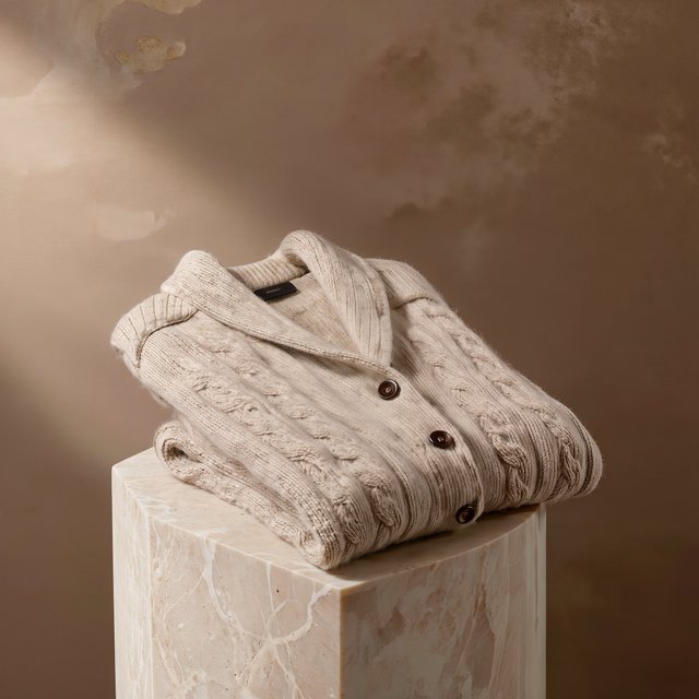 A folded unbranded merino cardigan rests on a honed limestone plinth, its chunky ribbed wool surface softly catching broad overhead diffusion and warm side bounce against a muted clay backdrop, with no tags, lettering, or graphics. — Kling v3