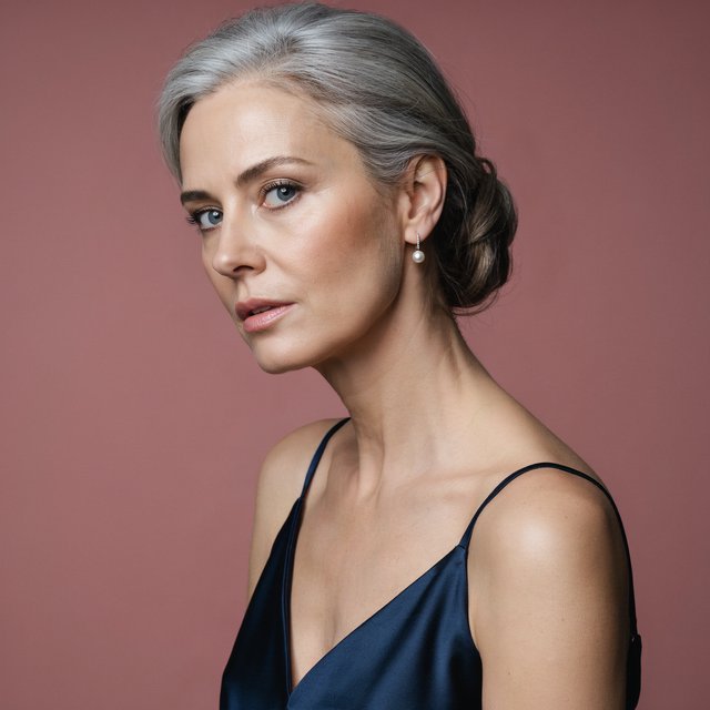 A silver-haired adult woman in a midnight-blue evening gown pauses with lipstick freshly applied, her composed gaze turned slightly aside; diffused rose studio light defines her face and shoulders, with restrained makeup and the polished finish of a fashion magazine. — Model 7